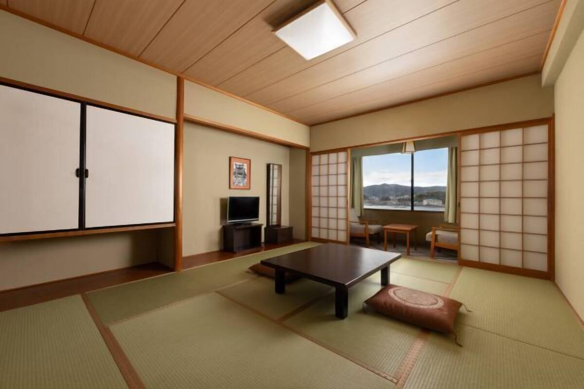 A traditional Japanese room with tatami flooring, a low table, sliding doors, a television, and a view of the outdoors.
