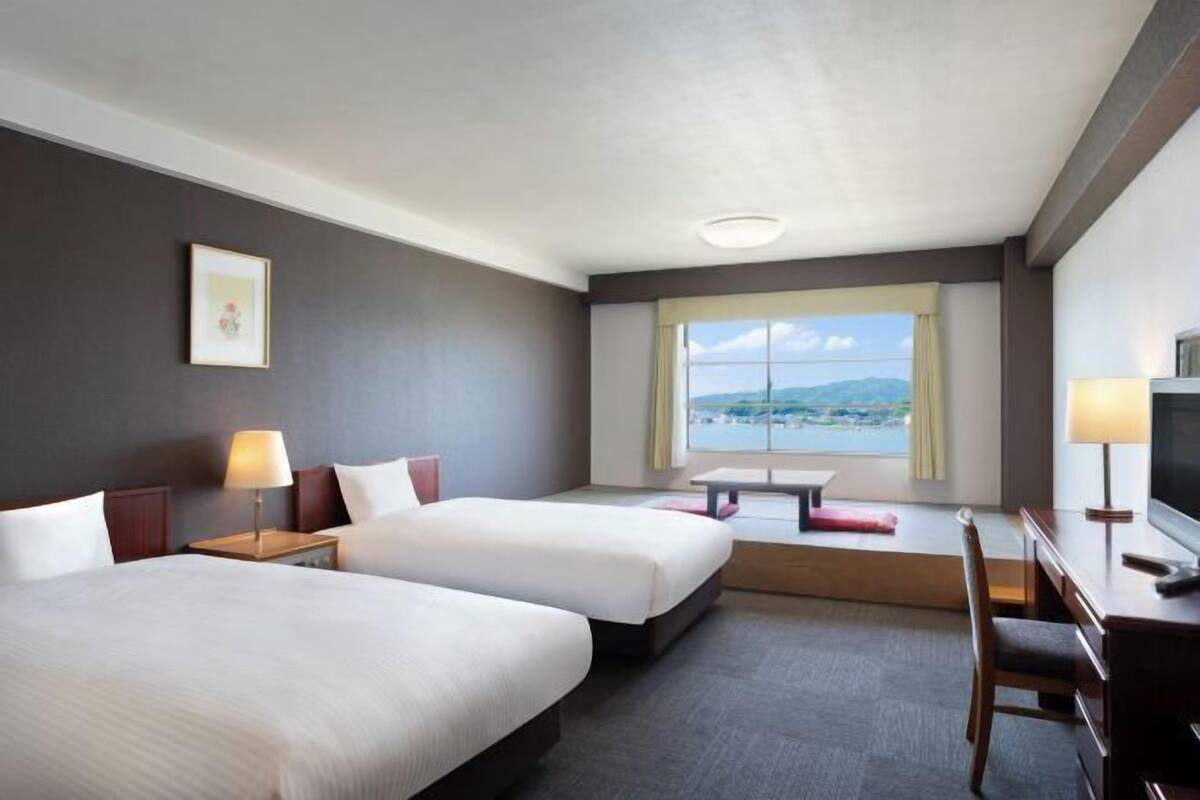 A hotel room with two beds, a desk, a chair, a TV, and a window with a view.