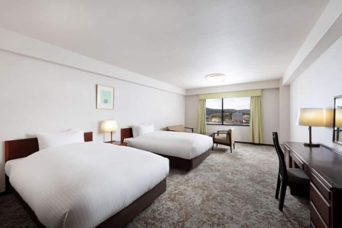 A hotel room with two beds, a desk, a chair, and a window with a view.