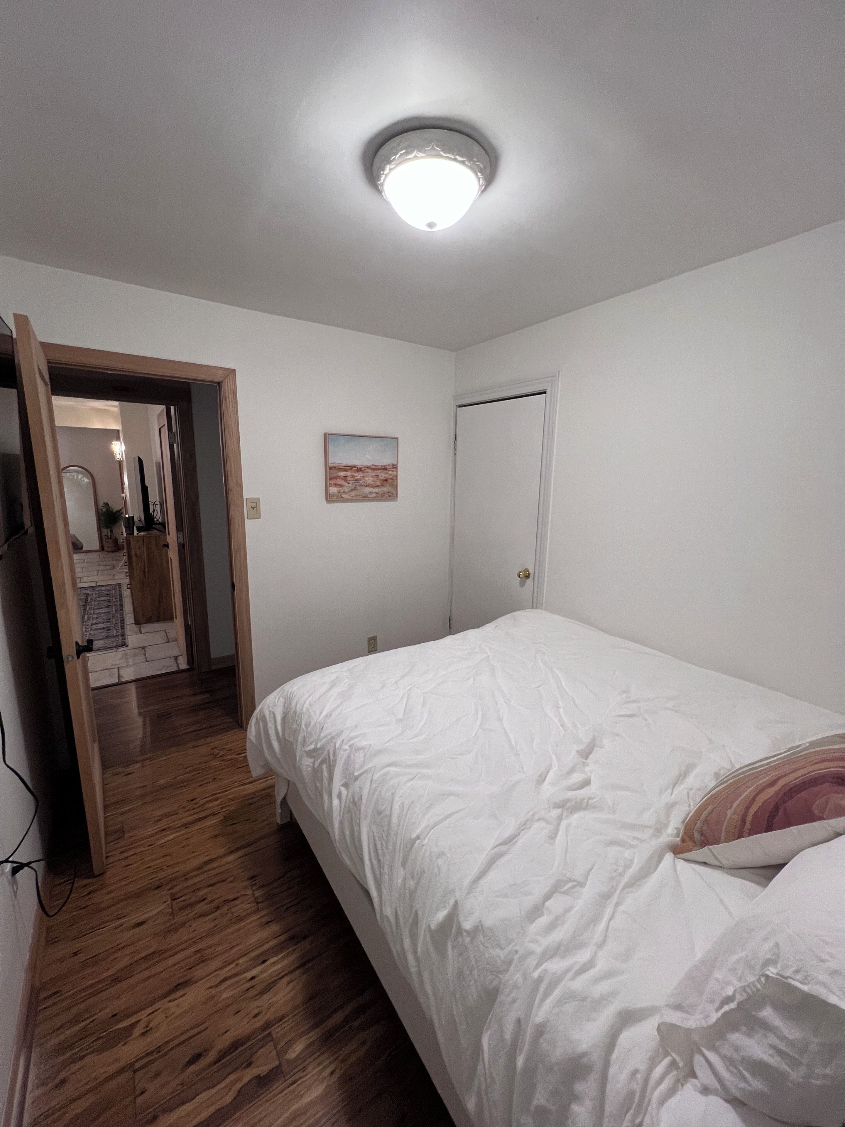 Ein Schlafzimmer mit Holzboden, einem weißen Bett, einer Tür und einer Deckenleuchte.