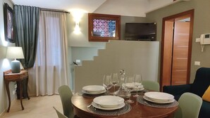 Un comedor con una mesa redonda de madera dispuesta para cuatro personas, rodeada de sillas verdes. Un televisor está montado en la pared sobre la mesa, y se ve una escalera al fondo.