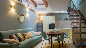 Una sala de estar acogedora con un sofá, una mesa de comedor redonda y sillas. Se ve una cocina al fondo con un refrigerador y una estufa.