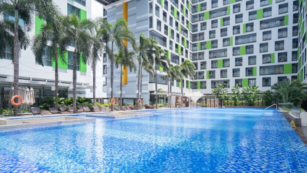 Una piscina con suelo de baldosas, rodeada de tumbonas y palmeras, con un edificio moderno al fondo.