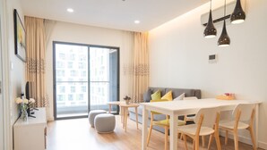 Una sala de estar moderna con sofá, mesa de comedor y sillas. Hay un ventanal con cortinas, una pequeña mesita de noche con un jarrón de flores, y un equipo de aire acondicionado instalado en la pared.