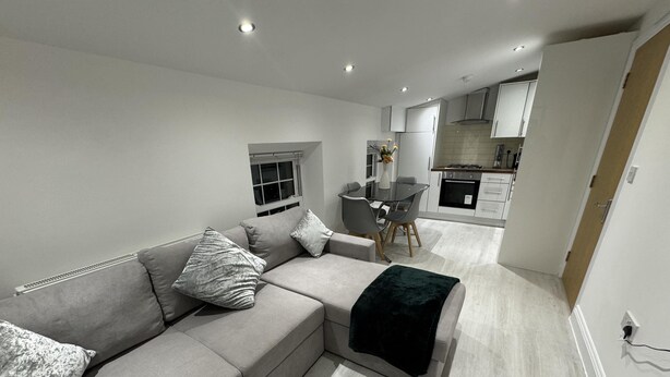 A modern living room with a grey sectional sofa, a glass dining table, and a kitchen in the background. - Beautiful 2 Bed Apartment in Walton on the Naze (Walton on the Naze)