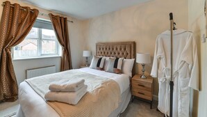 A neatly made bed with a tufted headboard, two bedside lamps, a wooden nightstand, and a robe hanging on a stand. - Charming 2-bed House in Middleton Manchester (Manchester)