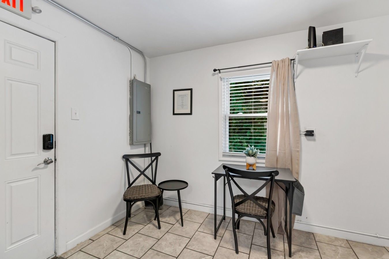 Une petite pièce propre, avec une porte blanche, une fenêtre avec des stores, deux chaises en osier, une petite table et une étagère avec quelques objets.