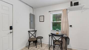 Une petite pièce propre, avec une porte blanche, une fenêtre avec des stores, deux chaises en osier, une petite table et une étagère avec quelques objets.