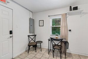 Una habitación pequeña y limpia, con una puerta blanca, una ventana con persianas, dos sillas de mimbre, una mesita y una estantería con objetos.