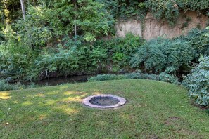 Un jardín ajardinado con un pozo de fuego circular, abundante follaje y un arroyo.