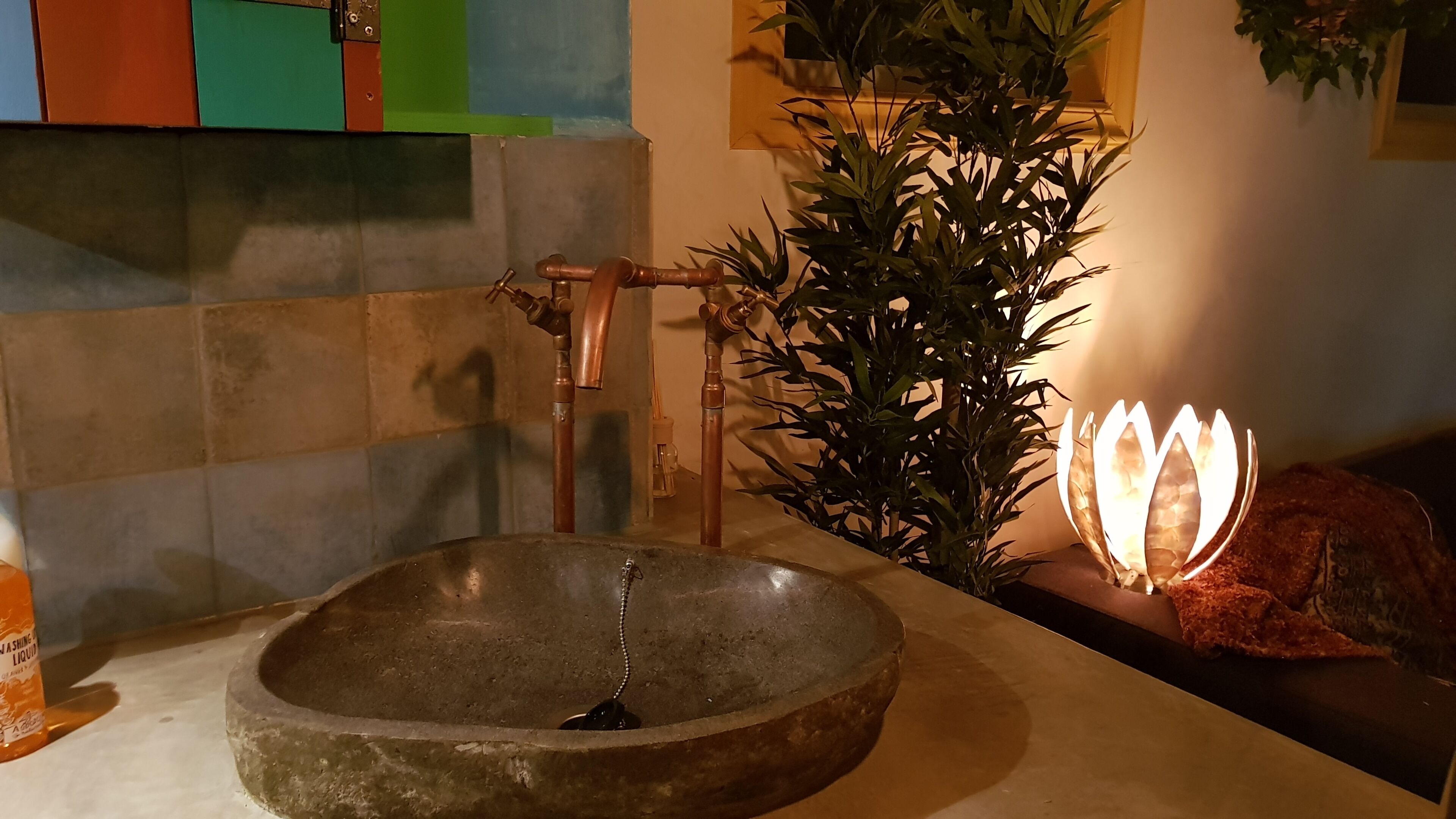Een badkamer met een wasbak van natuursteen, koperen leidingen, een plant in pot en een decoratieve lamp.