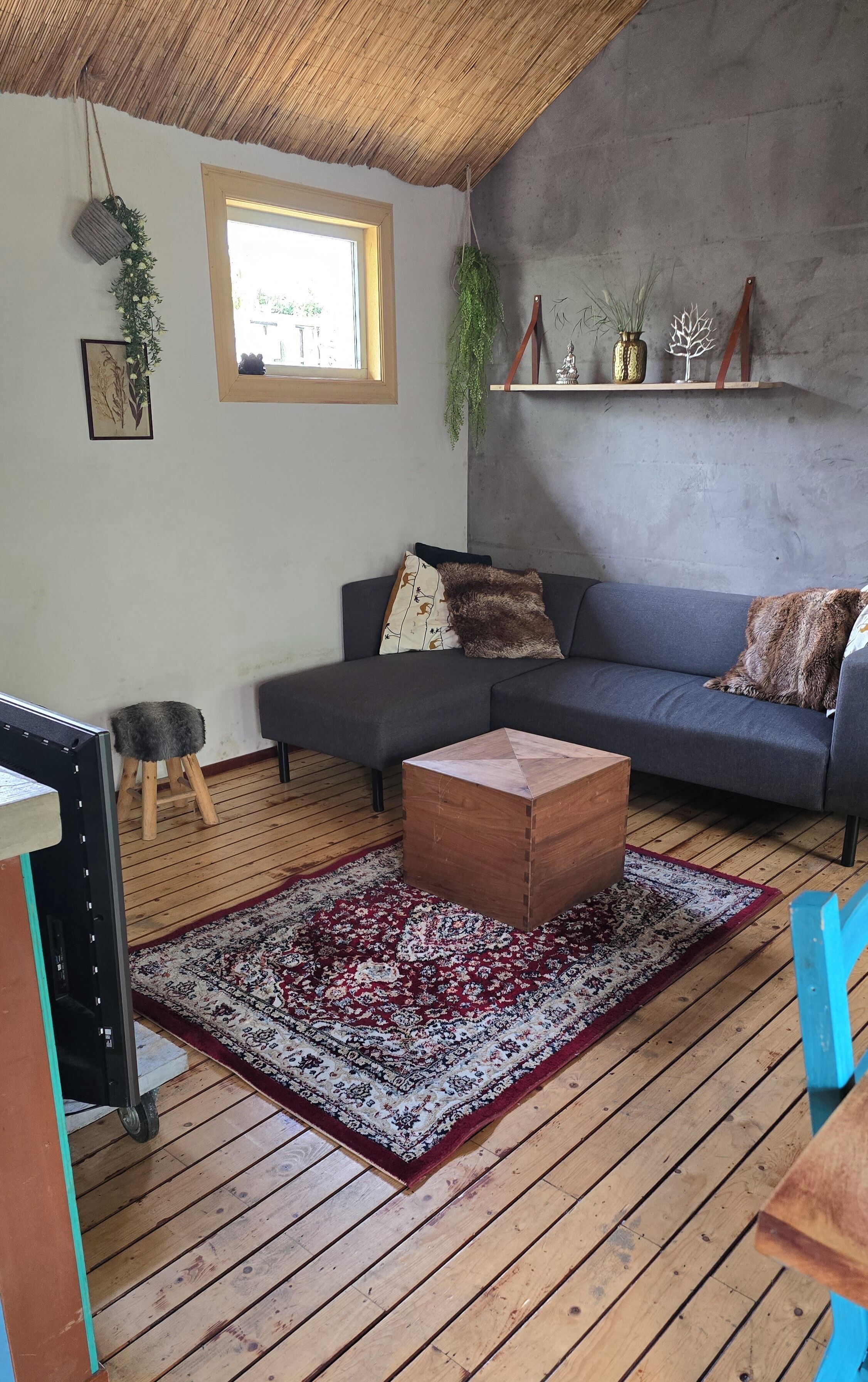 A living room with a grey sofa, a wooden coffee table, a red and white patterned rug, a small stool, and a window with natural light.