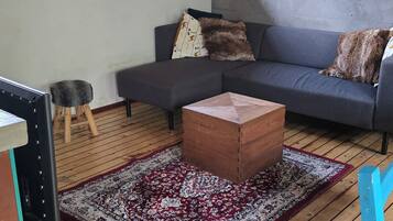 A living room with a grey sofa, a wooden coffee table, a red and white patterned rug, a small stool, and a window with natural light.