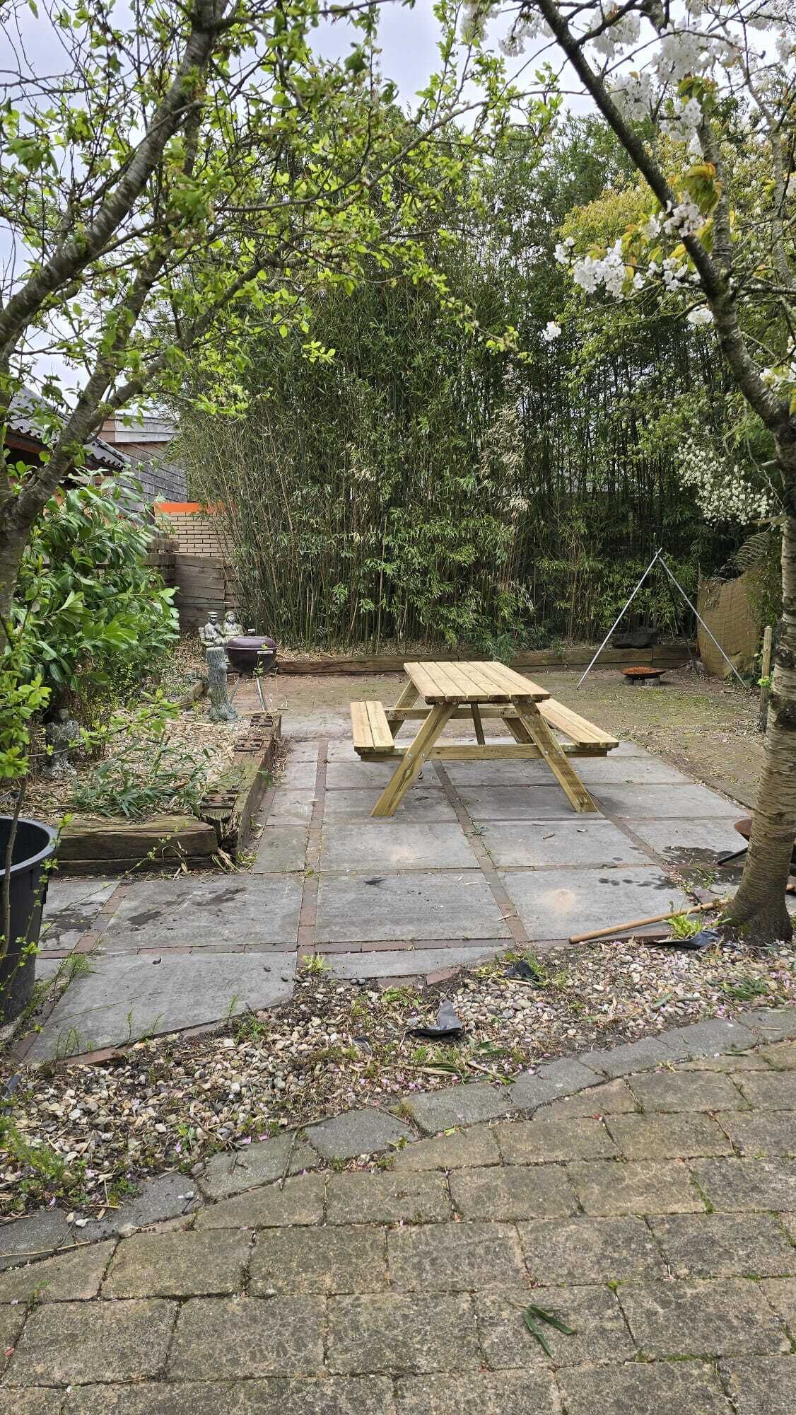 A wooden picnic table in a garden with stone paving, surrounded by dense foliage and a bamboo fence.