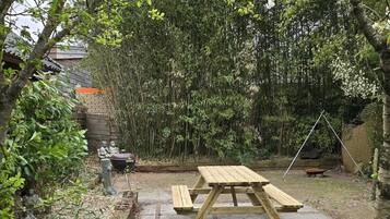 A wooden picnic table in a garden with stone paving, surrounded by dense foliage and a bamboo fence.