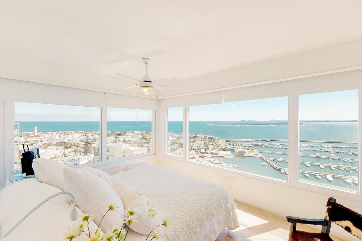 Une chambre avec un lit, une chaise et de grandes fenêtres donnant sur une marina et la ville.