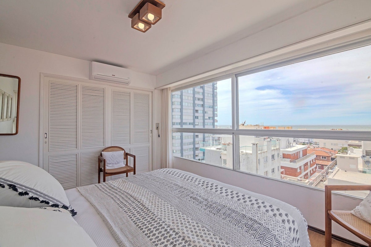 Une chambre avec une grande fenêtre, un lit, une chaise et une armoire.