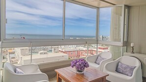 Ein Balkon mit Blick auf das Meer und Gebäude, weiße Stühle, ein Holztisch und eine Vase mit violetten Blumen.