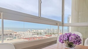 Ein Balkon mit Blick auf Stadt und Meer, ein Holztisch und eine Vase mit Blumen.