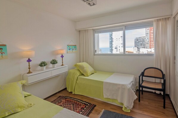 A bedroom with a bed, a chair, a window with a view of buildings, and a small shelf with decorative items.