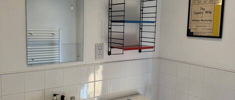 A bathroom with a white sink, toilet, and mirror. There is a shelf above the toilet and a framed poster on the wall.