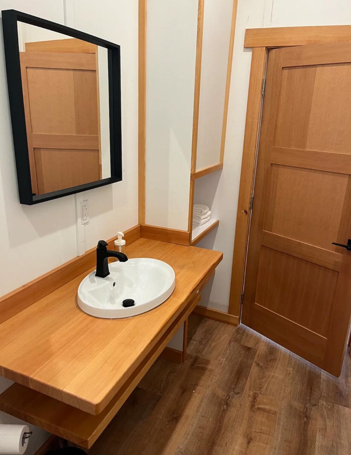 Une salle de bain avec un meuble-lavabo en bois, un lavabo blanc, un robinet noir, un miroir et une porte en bois.