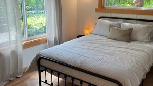 A bedroom with a bed, a window with curtains, a bedside lamp, and a view of greenery outside.