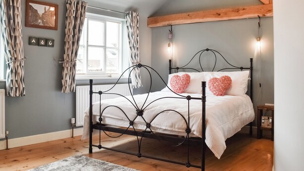 Une chambre avec un cadre de lit en métal, du linge de lit blanc, des coussins en forme de cœur, un plancher en bois, une fenêtre avec des rideaux et un tableau accroché au mur.