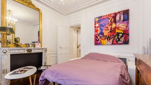 A bedroom with a bed, a dresser, a vanity with a mirror, a chandelier, and a colorful artwork on the wall. - Luxury Près des Champs Elysées (Paris)