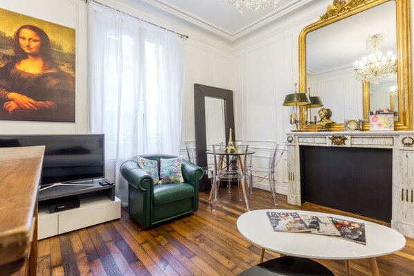 Un salon avec un téléviseur à écran plat, un fauteuil vert, une petite table ronde et un grand miroir.