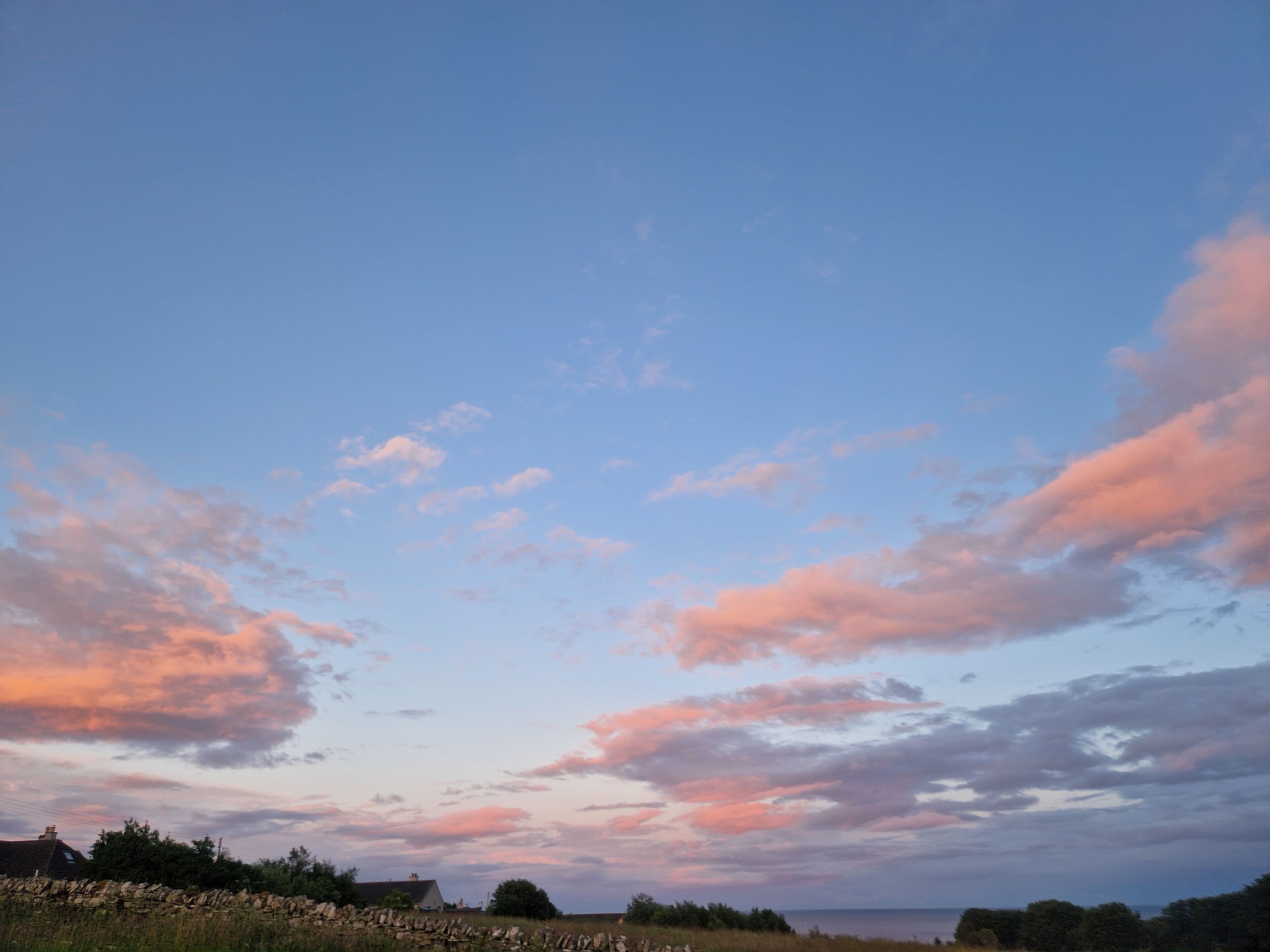 A rural landscape with a stone wall, a house, and a clear sky with clouds.