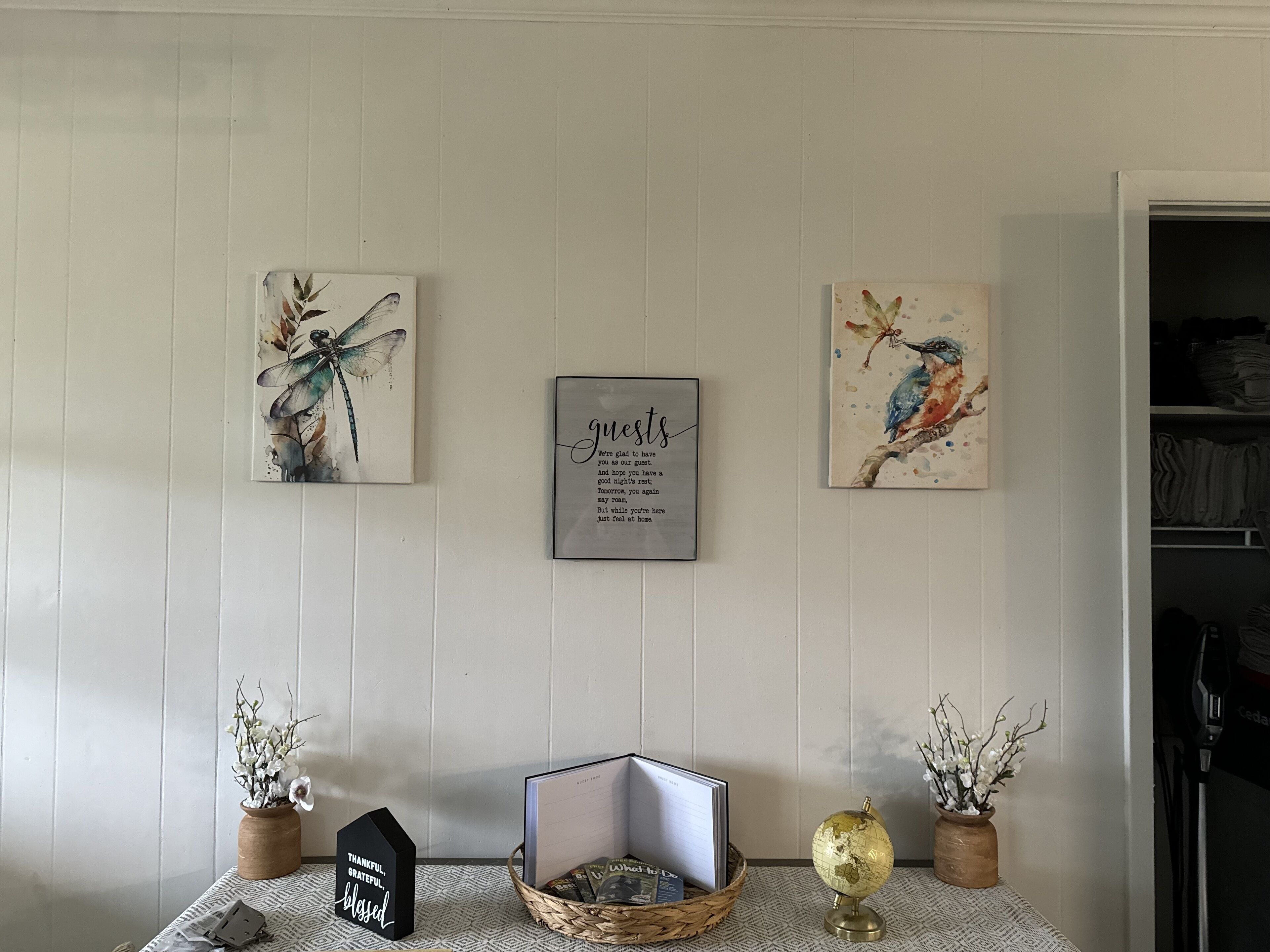 Una habitación con una pared blanca con paneles, una cita enmarcada y dos obras de arte enmarcadas que representan una libélula y un pájaro. Hay una pequeña mesa con un globo terráqueo, una cesta y un letrero que dice "THANKFUL, GRATEFUL, blessed".