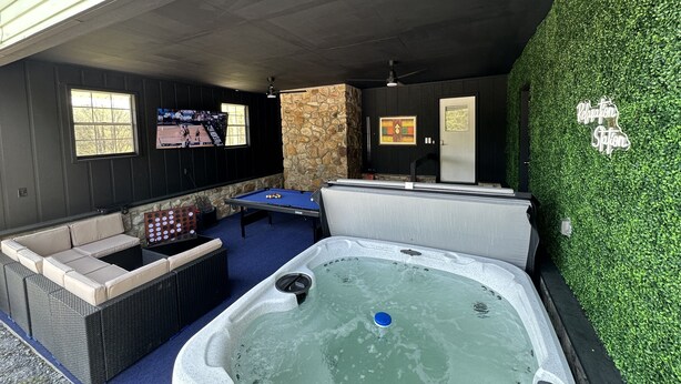 Une table de billard, un jacuzzi et une table de jeu de fléchettes dans une pièce aux murs en pierre et bordée d’une haie verte.