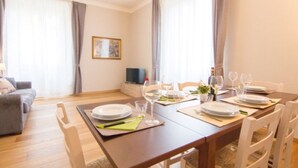 A dining area with a wooden table set for four, white chairs, and a television on a stand in the background.