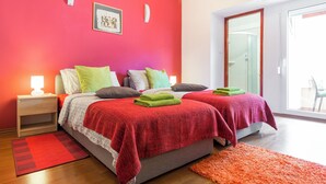 Un dormitorio con dos camas, una mesita de noche con lámpara, una pared roja y un ventanal grande.