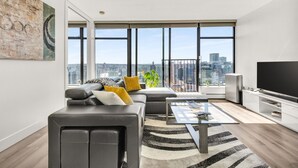Un salón moderno con sofá, mesa de centro y televisor. Amplias ventanas ofrecen vista a la ciudad.