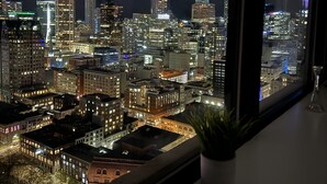 Un paisaje urbano nocturno visto a través de una ventana, con edificios iluminados y una pequeña planta en maceta en el alféizar.
