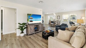 A spacious living room with a large flat-screen TV, a wooden entertainment unit, a beige sofa, and a dining area with a table and chairs.