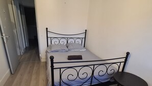 Habitació petita amb un llit individual de vambes metàl·liques, una taula petita i una porta que dona a una altra habitació.