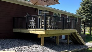 A wooden deck with a railing, a pergola with an umbrella, and a brick building in the background. - Country side retreat, perfect place for family fun and hunting. (Marshall)