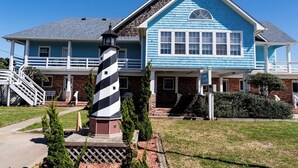 A two-story house with a blue exterior, a white porch, and a black and white striped lighthouse. - Hugo - Offshore Beach Club (Buxton)