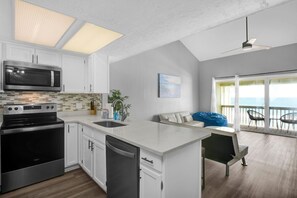 A modern kitchen with a built-in microwave, stove, and dishwasher. There is a large island with a sink, and a dining area with a table and chairs. The kitchen has a large window and a view of the ocean.