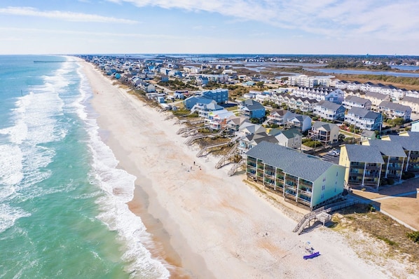 An aerial view of a coastal residential area with beachfront properties, a pier extending into the ocean, and a clear sky.