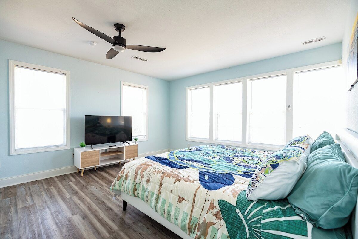 Ein Schlafzimmer mit einem Bett, einem Fernseher, einem Deckenventilator und großen Fenstern.