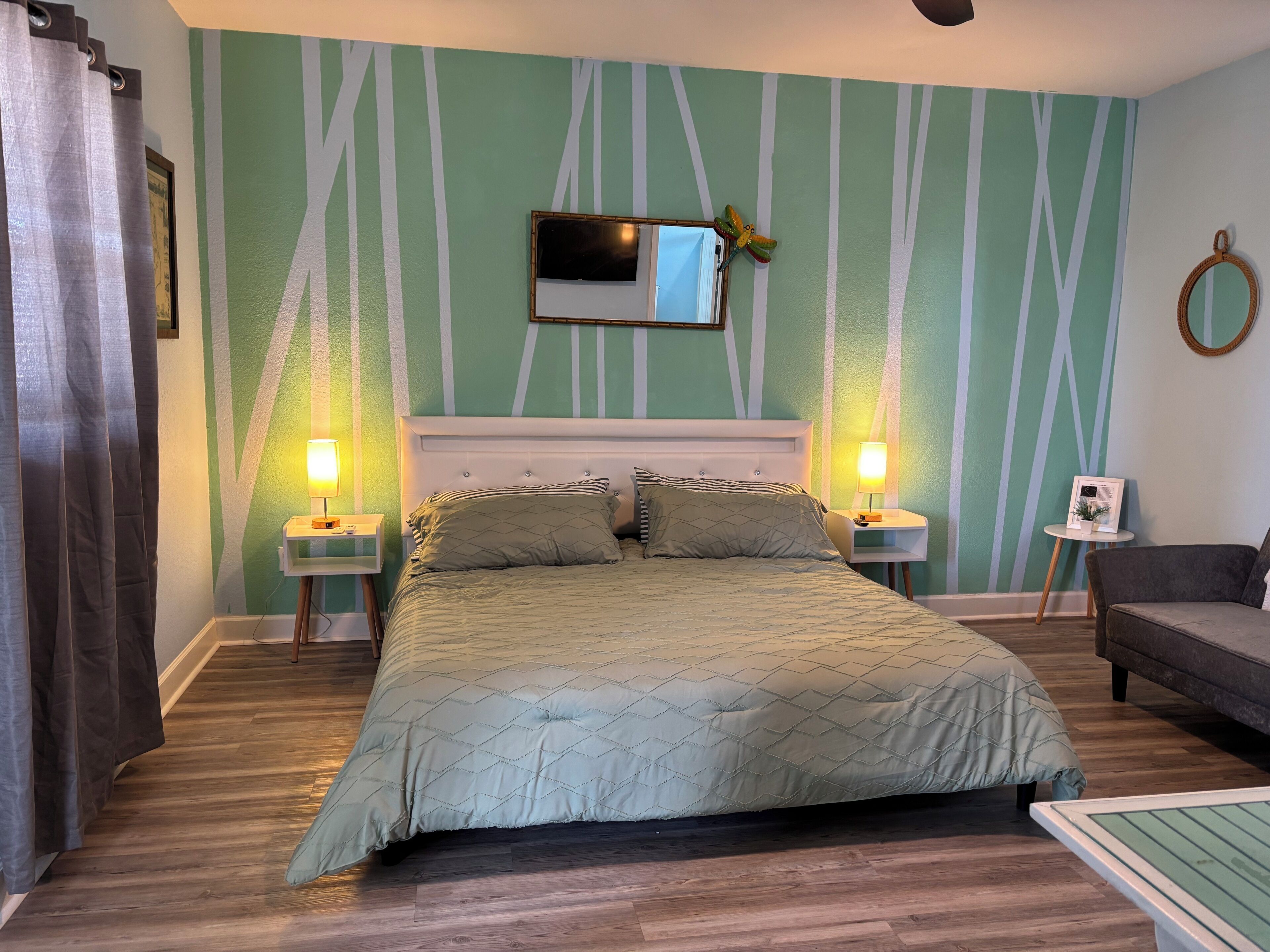 A bedroom with a bed, two bedside tables with lamps, a mirror, a small side table, a couch, and a wall with a striped pattern.