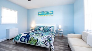 Une chambre à coucher avec un lit, un ventilateur de plafond, une table de chevet, une commode, une fenêtre et un tableau au mur.