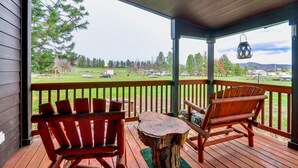 Une terrasse en bois avec deux fauteuils Adirondack et une petite table, donnant sur une zone herbeuse bordée d’arbres et où l’on aperçoit des véhicules au loin.