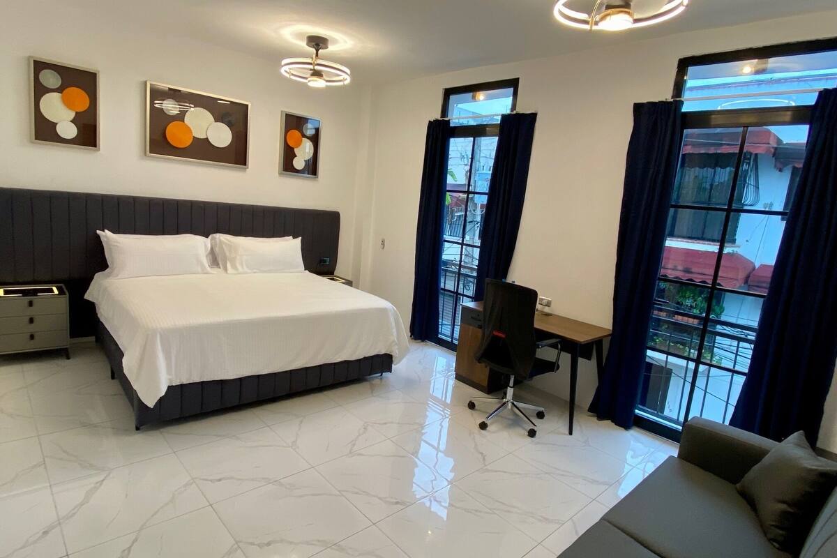 A modern hotel room with a large bed, a desk, and a sofa. There are three wall art pieces above the bed and a large window with curtains.