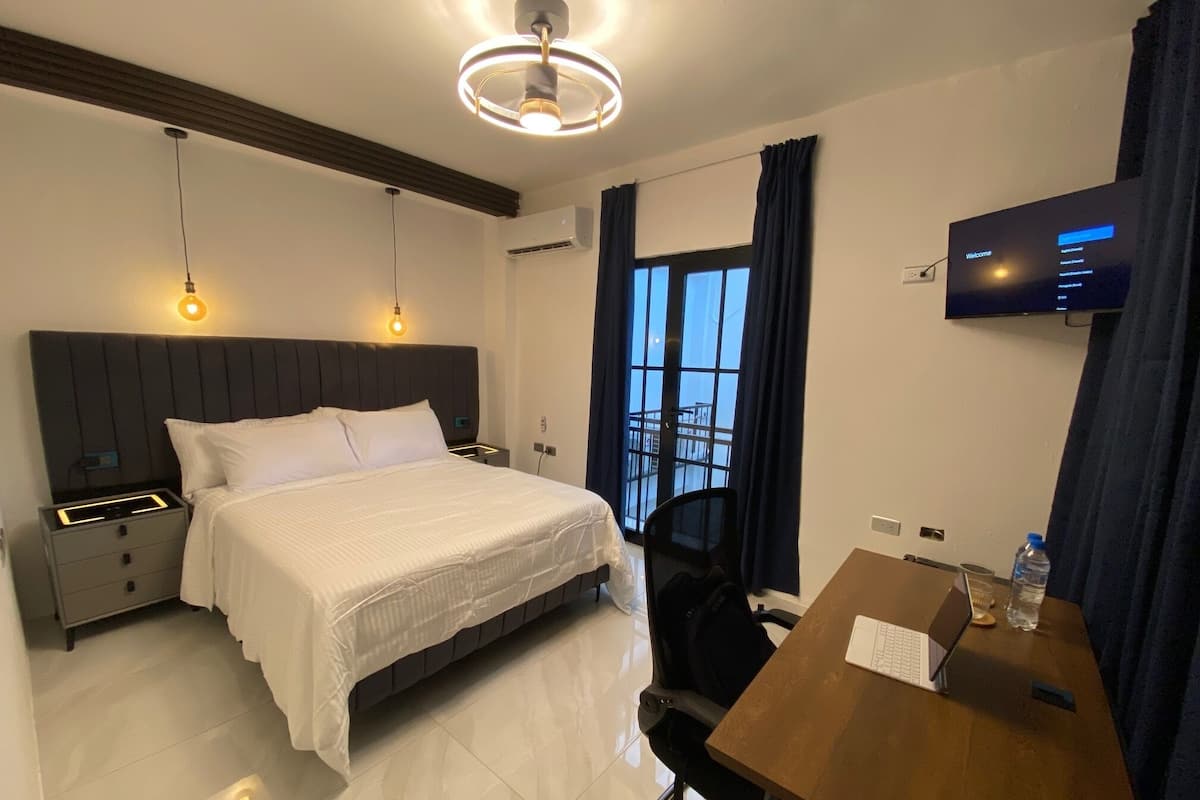 A hotel room with a bed, bedside tables, a desk with a laptop, a chair, a TV, and a window with curtains.