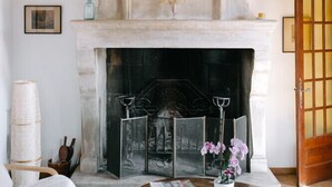 A room with a fireplace, wooden beams, a white wall, a framed picture, a table with a vase of flowers, and a door with a grid pattern. - Mas in Gordes with swimming pool (Gordes)